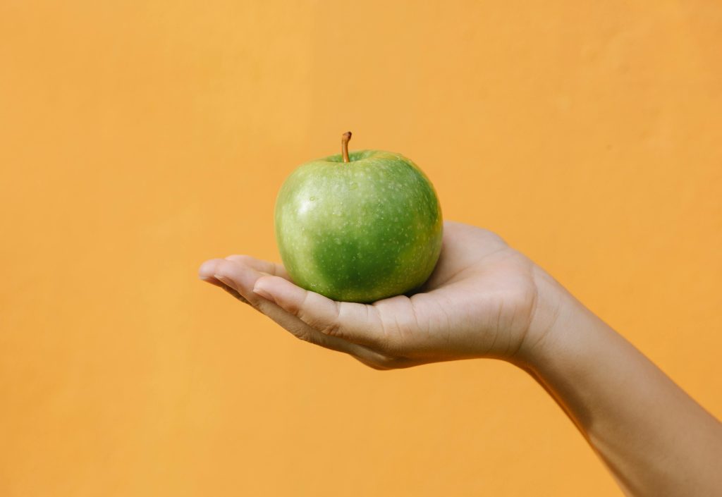Une pomme verte tenue par la pomme de main droite tournée vers le haut sur un fond d'écran jaune désaturé.