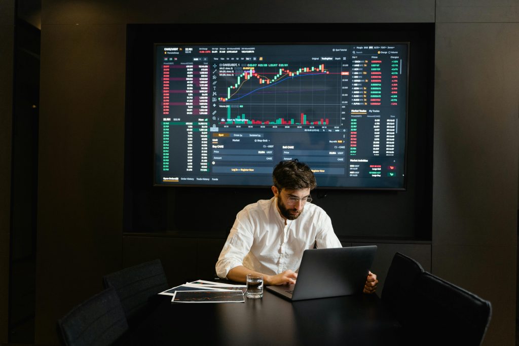 Un homme travaille à son bureau avec pour arrière-plan des écrans qui affichent les les graphiques et les données du marché boursier.