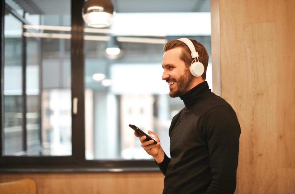 Un homme debout, portant les écouteurs de couleur blanc avec son smartphone à la main. Souriant, il semble entrain d'avoir une conversation agréable. Le tout dans un cadre éclairé d'un antichambre de bureau.