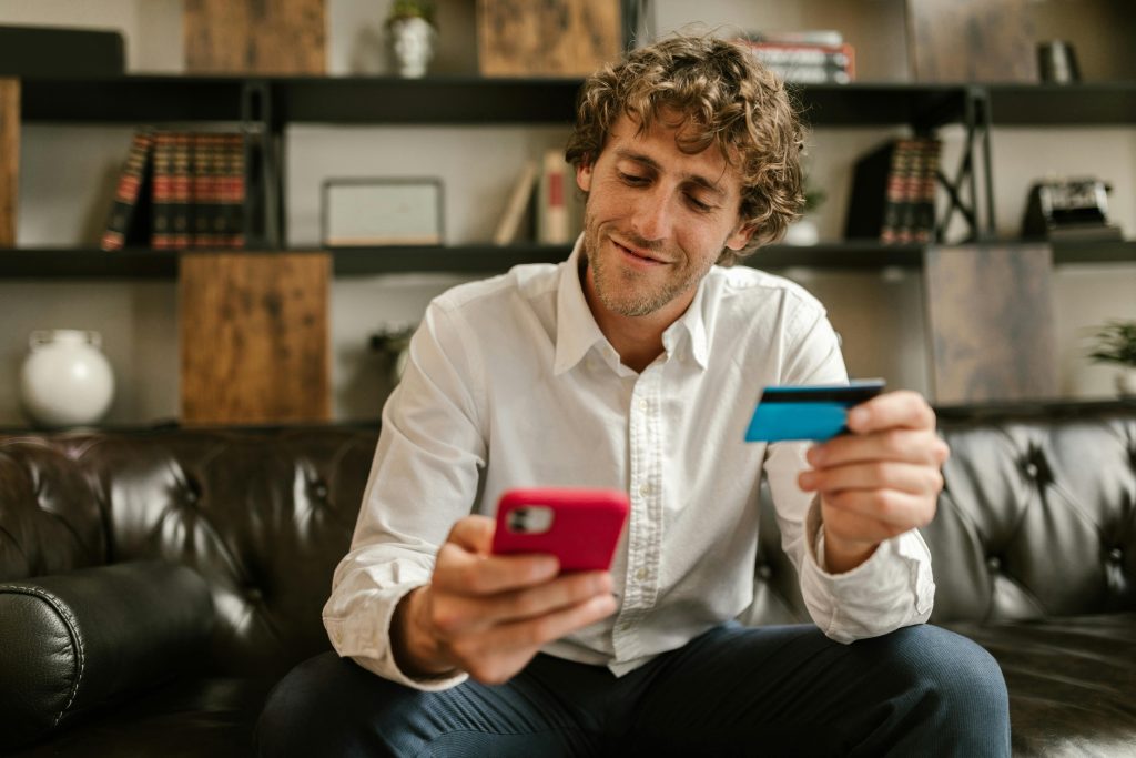 Un homme assis dans un canapé avec un smartphone de couleur rouge dans sa main droite, et une carte bancaire bleue dans sa main gauche. Il regarde l'écran de son téléphone avec un sourire satisfait.