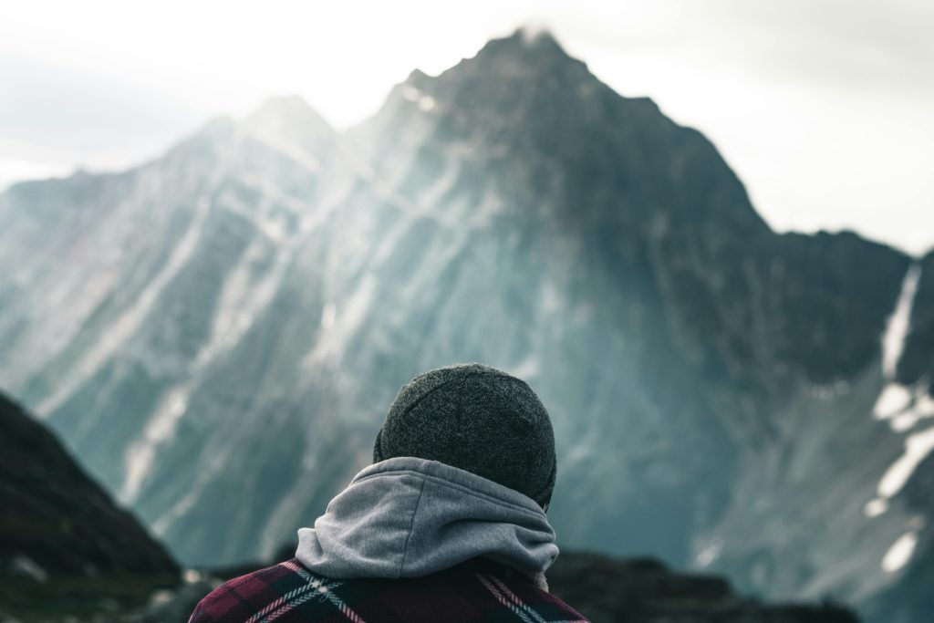 Photo prise de dos d'une personne qui se trouve face à une montagne à gravir. La scène est bercée par un prisme solaire venant du coin haut à gauche.